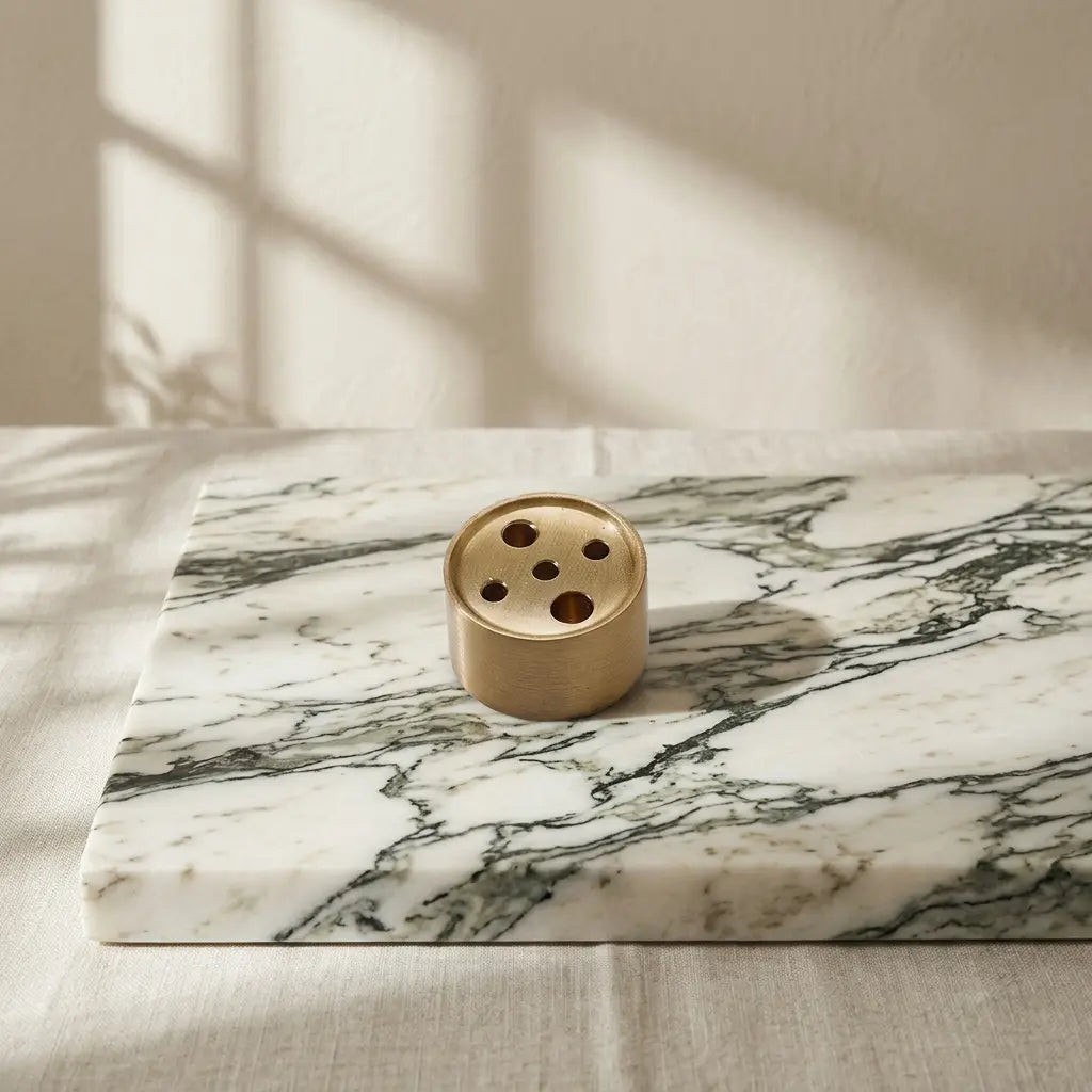 Solid brass cylinder incense holder with multi-size holes displayed on a premium Arabescato marble plinth.