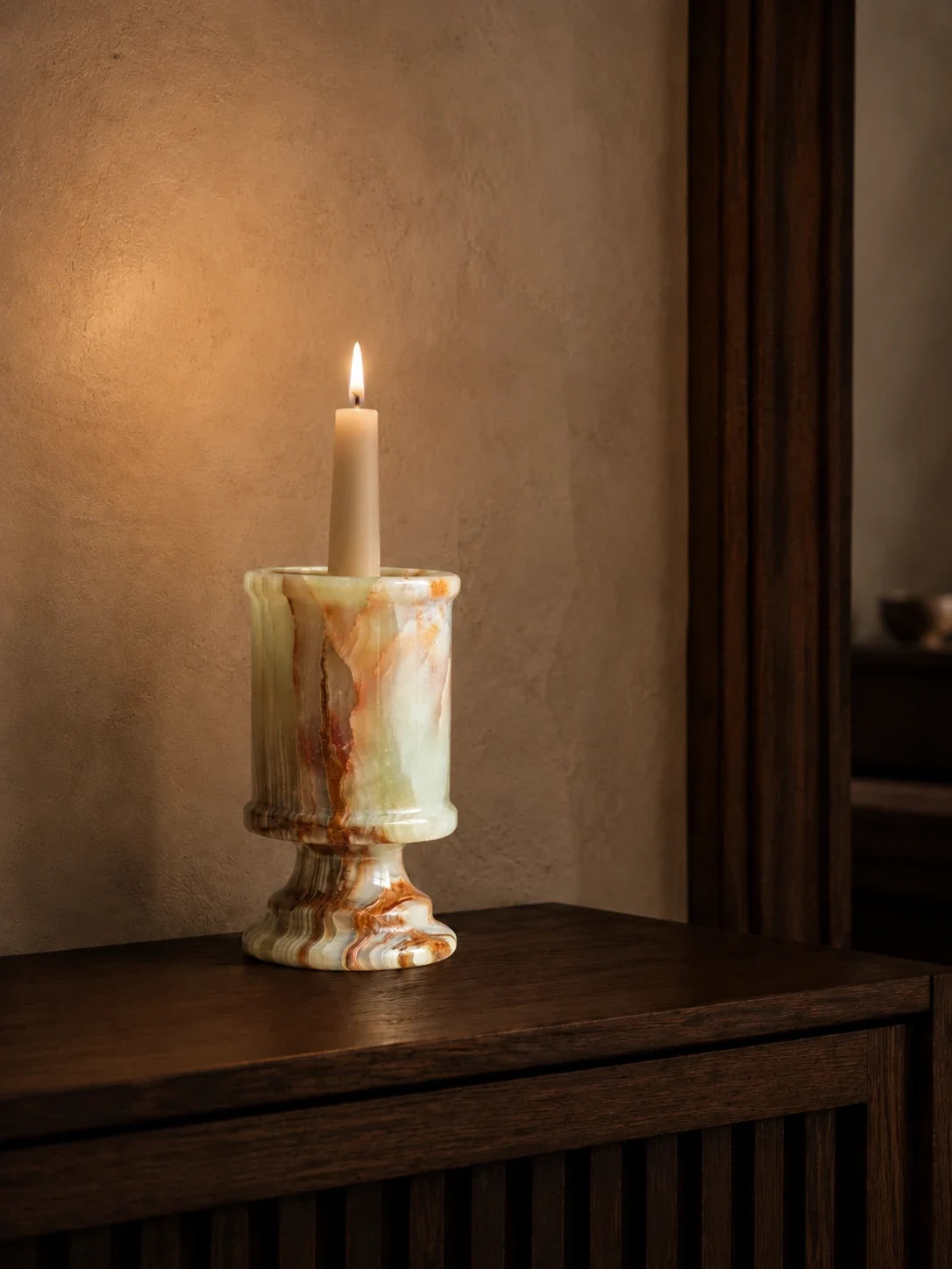 Natural onyx candle holder with a lit ivory candle on a dark walnut console, revealing layered green, amber, and brown mineral patterns in warm evening light.