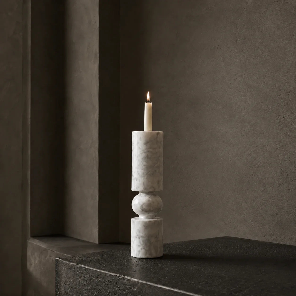 White marble sculptural candlestick with a lit ivory taper candle in a dark architectural gallery interior
