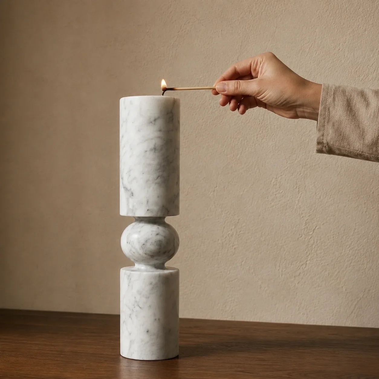 White natural marble pillar candle holder with a sculptural stacked design being lit by hand on a walnut table, showing its realistic scale, rounded stone sphere detail, grey veining, and polished finish.