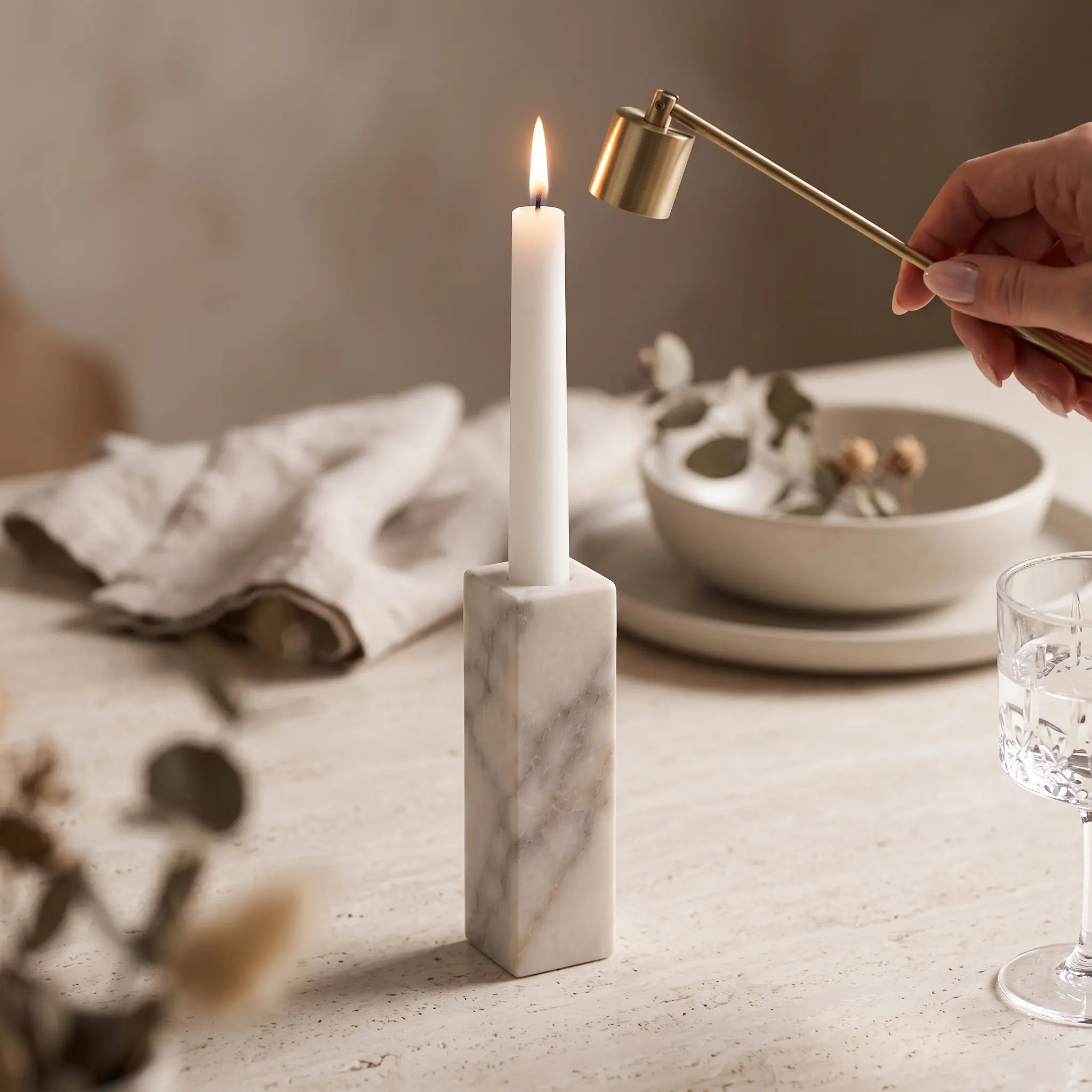 Monolith Candle Holder in White Marble styled on a dining table with a lit taper candle and brass snuffer