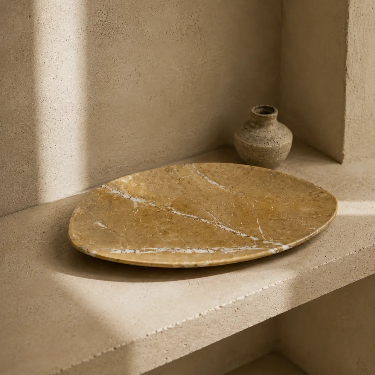 Aged golden marble leaf-shaped tray displayed on a limestone shelf with a ceramic vessel, highlighting its natural mineral veining, organic silhouette, and polished stone surface.