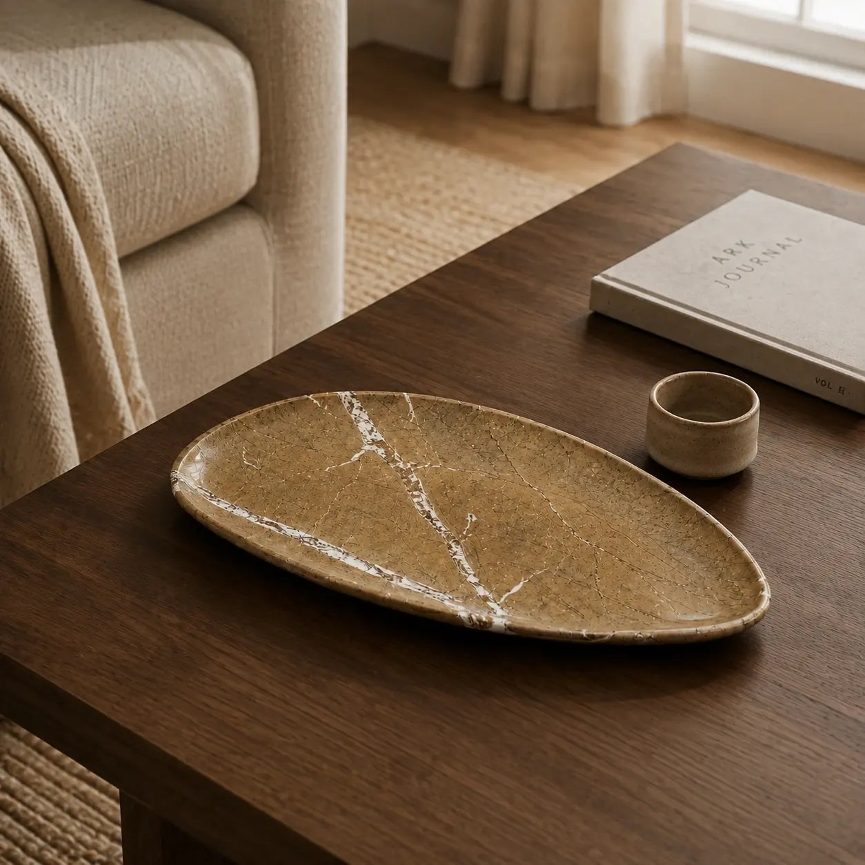 Aged golden marble leaf-shaped tray on a walnut table with a ceramic vessel and art book, showcasing its natural mineral veining, organic silhouette, and polished stone surface.