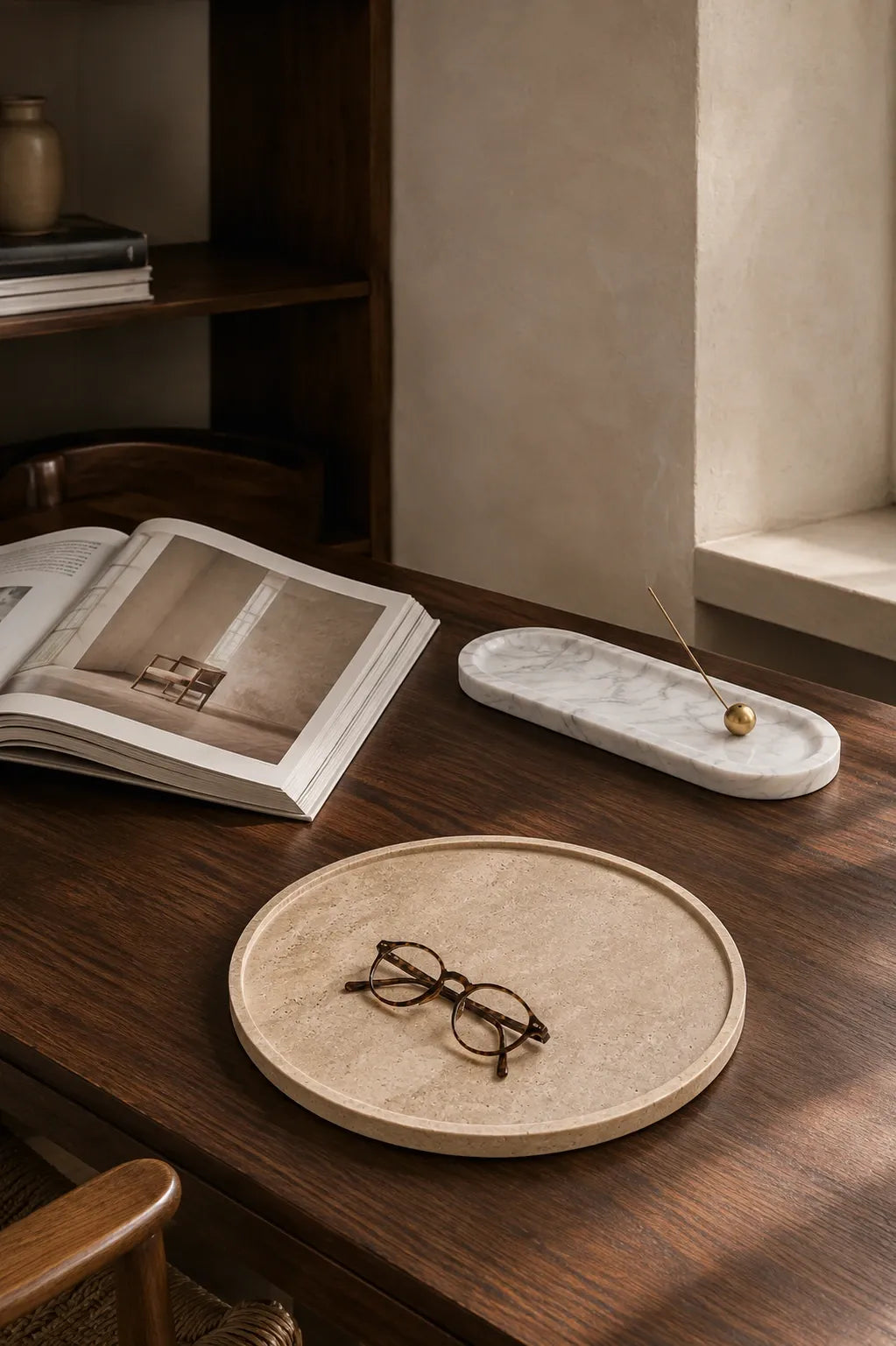 Travertine round tray with reading glasses beside a marble incense tray and open architecture book on a dark walnut desk.