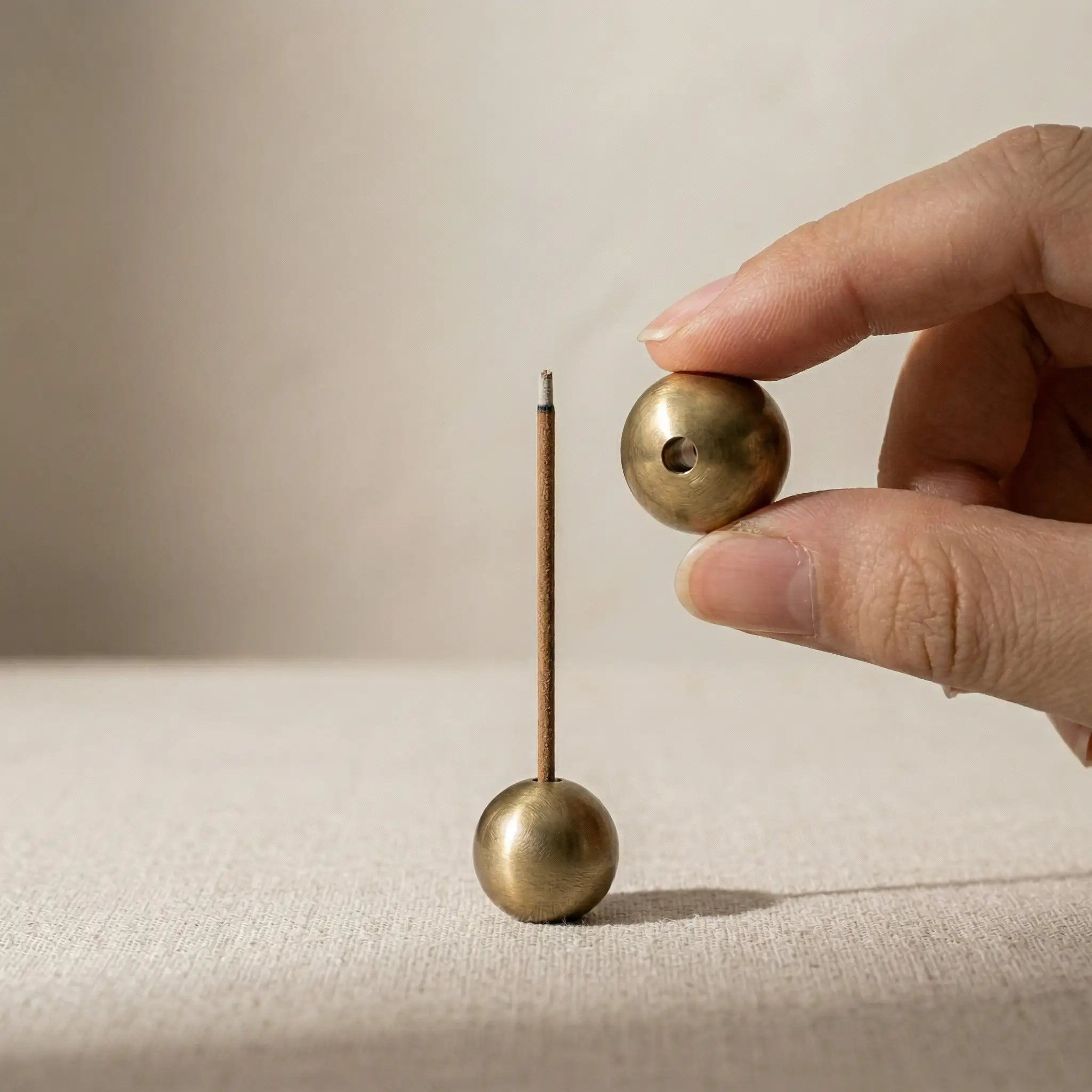 A person's hand placing a Brass Sphere Relic onto an incense stick to demonstrate its minimalist design and scale.