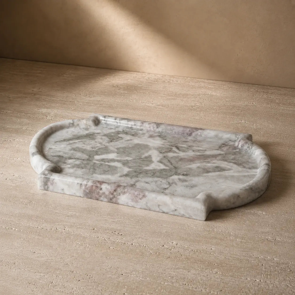Grey and white natural marble tray with an organic rounded rectangular shape, polished stone edge, and unique mineral veining displayed on a warm limestone surface.