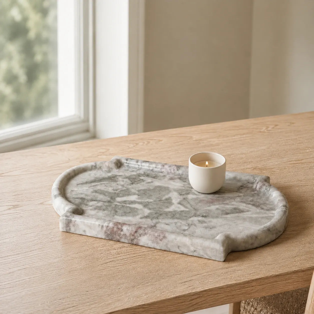 Natural marble tray with grey, white, and subtle pink veining, styled with a small ceramic candle on a light oak table in soft morning light.