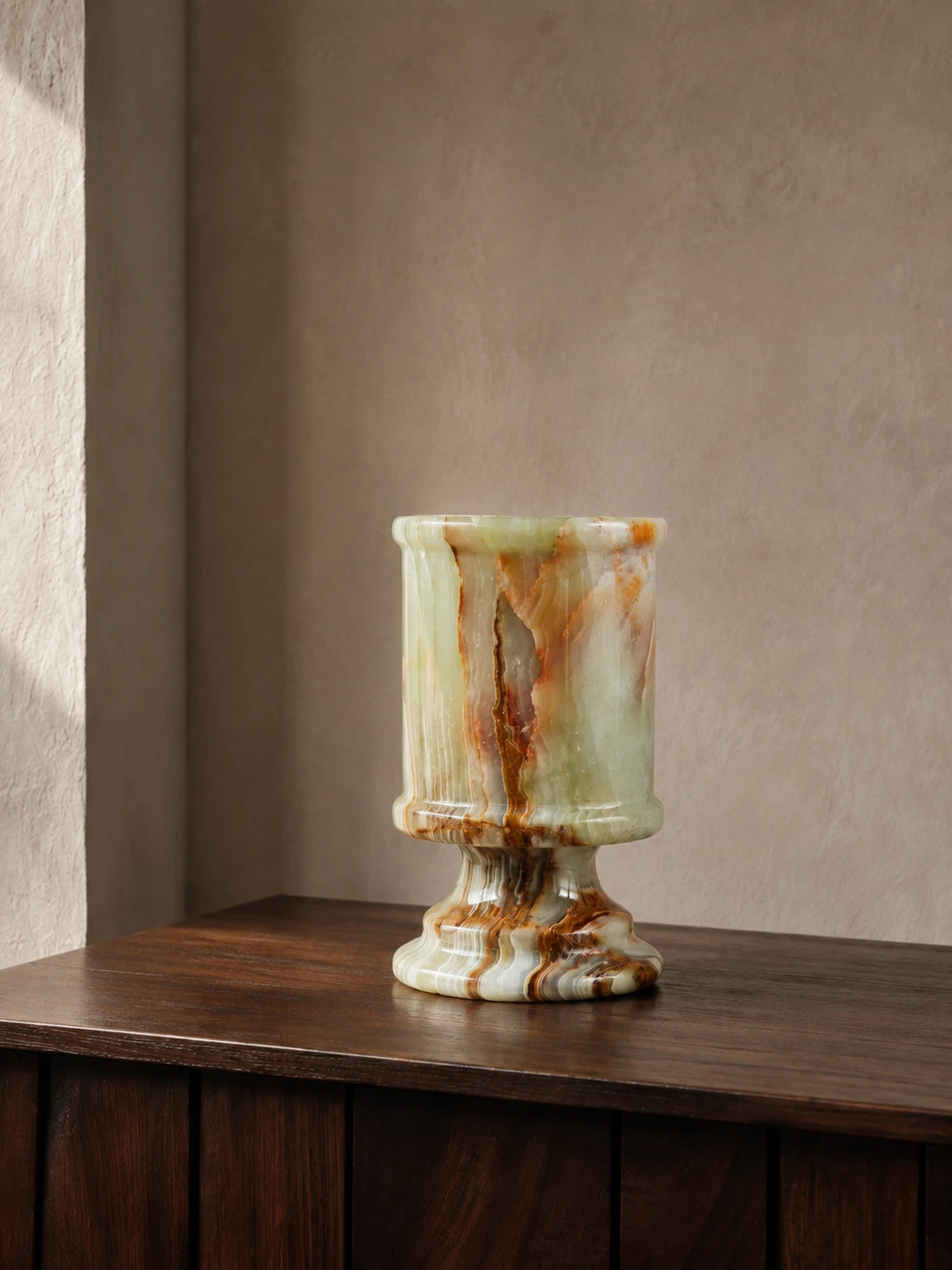 Natural onyx candle holder displayed on a dark walnut console in a quiet architectural interior, featuring layered green, amber, and brown mineral patterns with a polished stone finish.