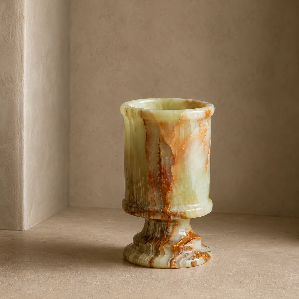 Natural onyx marble candle holder with a sculptural cylindrical form, polished stone finish, and layered green, amber, and brown mineral patterns displayed in a warm architectural interior.