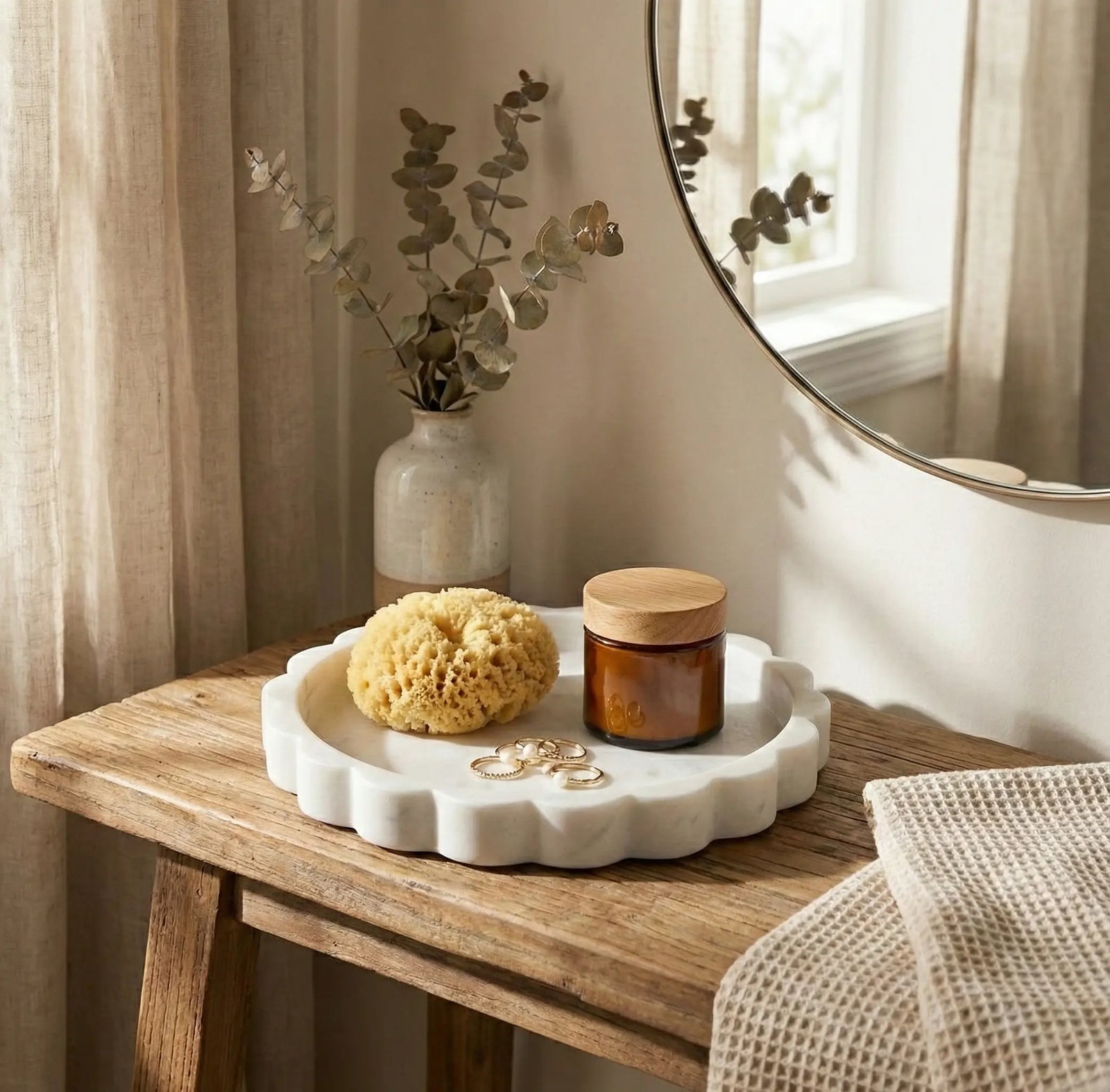 White scalloped marble circular tray styled on a wooden vanity with a candle, sponge, and bathroom accessories.