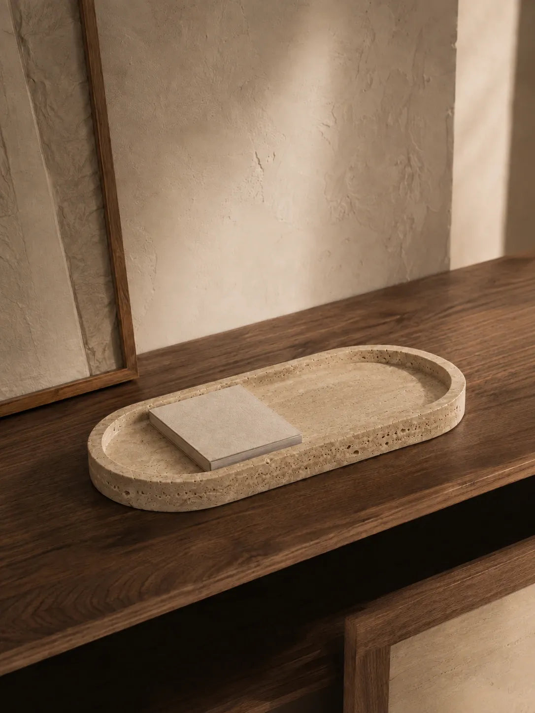 Natural travertine long tray styled on a dark walnut table with a simple book, showing the warm stone texture, elongated silhouette, and natural mineral variation in afternoon light.