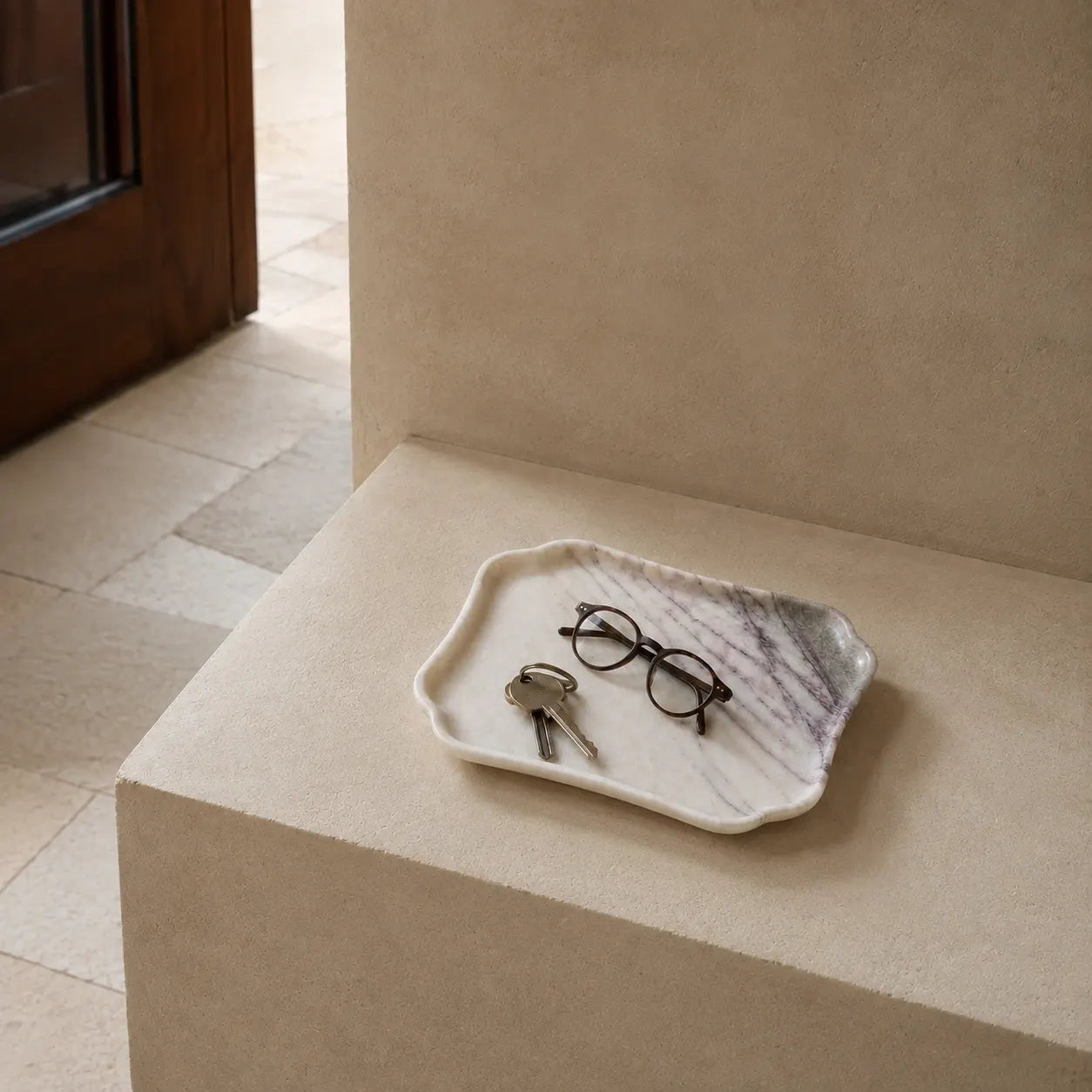 White marble catchall tray on a limestone entryway surface with keys and glasses, showcasing its scalloped edge, natural stone texture, and compact everyday scale.