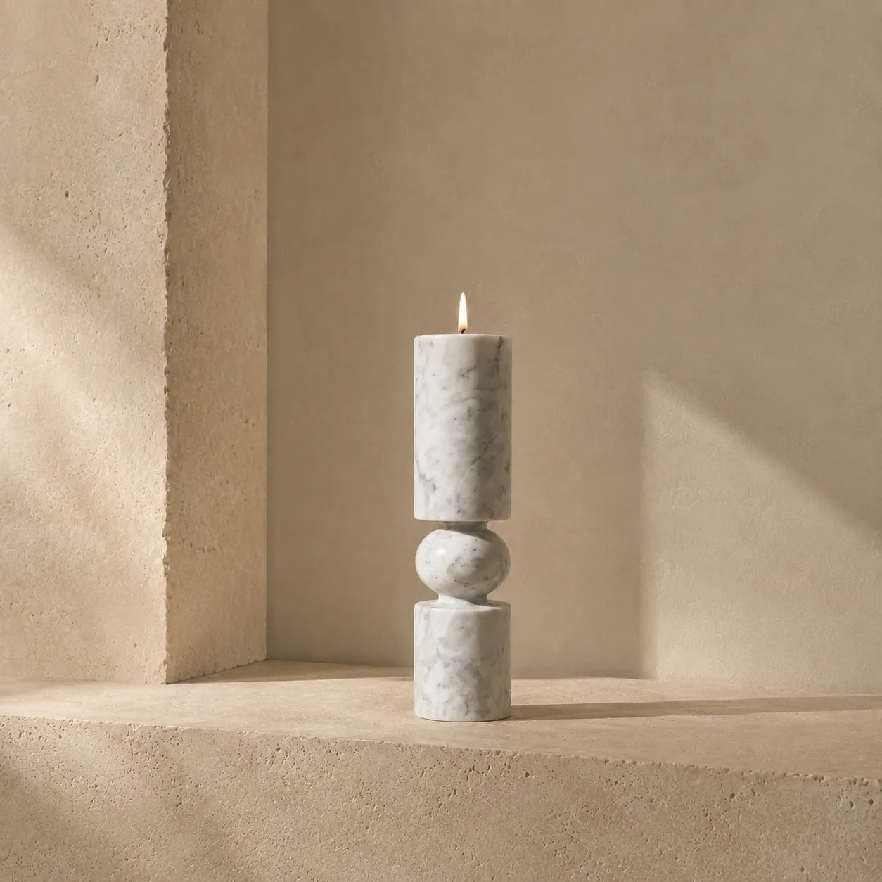 White natural marble pillar candle holder with a sculptural stacked design displayed on a limestone shelf in an architectural interior, featuring grey veining, a rounded stone sphere detail, and polished marble finish.