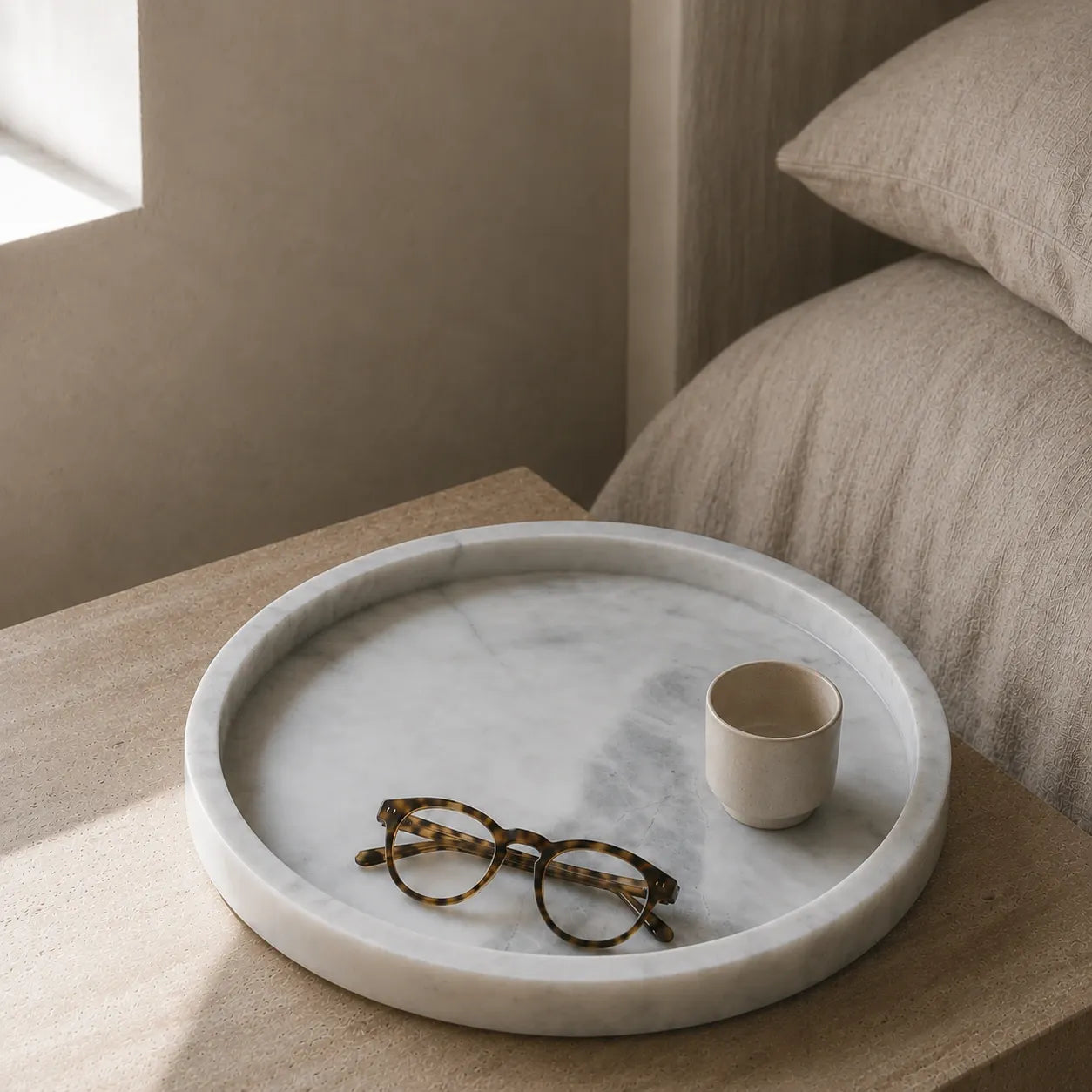 White natural marble round tray styled with reading glasses and a ceramic cup beside the bed, highlighting its 3 cm thick polished edge, subtle grey veining, and timeless carved stone design.