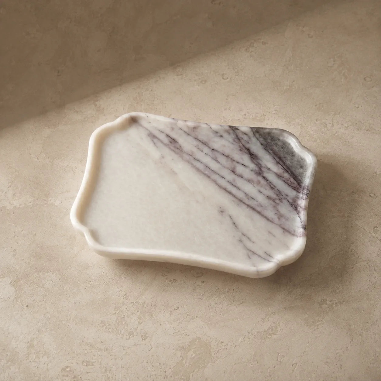 White scalloped natural marble tray on a limestone surface, showcasing its sculptural edge, polished finish, and unique grey-purple stone veining.